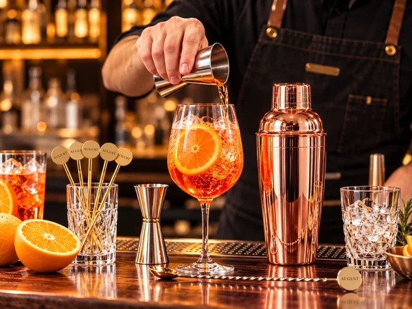 Bartender preparing an orange Spritz cocktail beside a rose gold cocktail shaker and bar tools